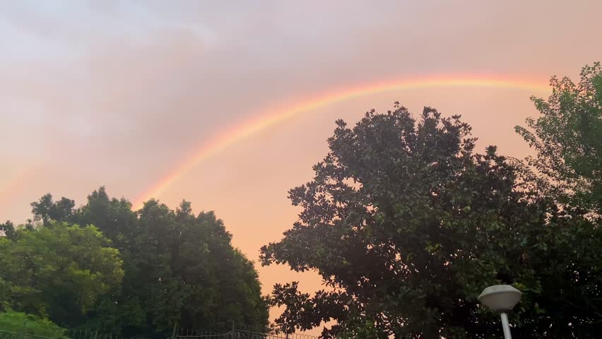 Soft sunset sky with a vivid single rainbow arching above leafy green trees in Islamabad, Pakistan, creating a warm natural scene after rain.