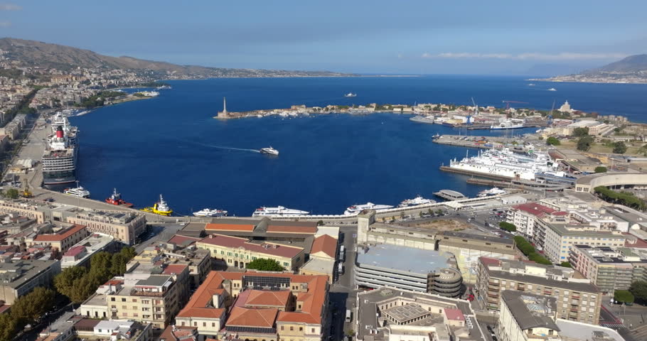 Aerial view of the city port of Messina, Sicily, Italy. Panorama of a crescent-shaped harbour. Large cruise ships and various vessels are docked along the quay. The historic center is in foreground.