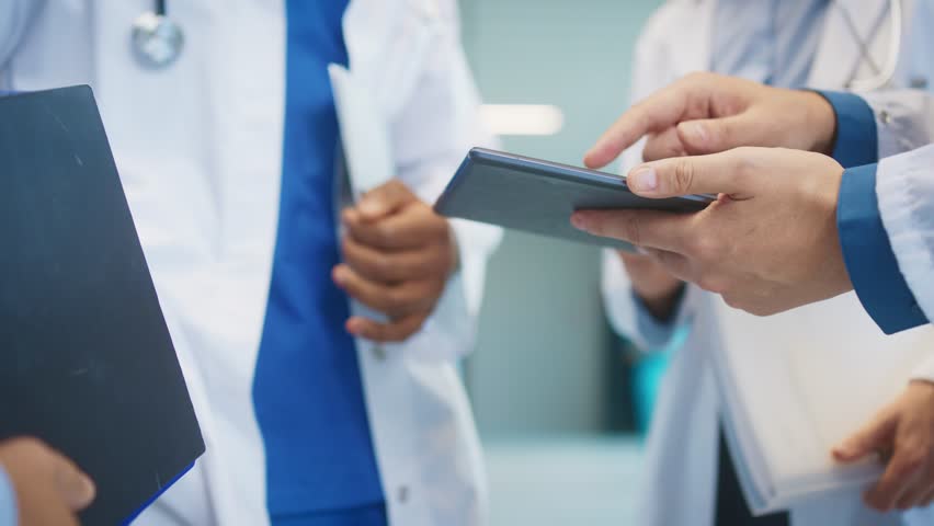 Close view of medical staff holding tablet in bright hospital corridor. Checking patient data. Touching screen with fingers and reviewing digital notes while preparing next clinical action.