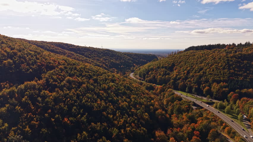 Golden autumn leaves blanket the rolling hills a winding road weaves through the lush valley and distant peaks rise towards a bright sky filled with soft clouds.