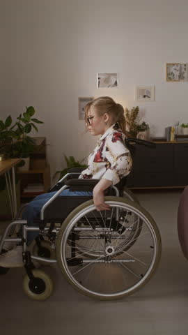 Vertical side view of Caucasian young wheelchair user pushing herself to wooden desk and typing on laptop while spending time at home