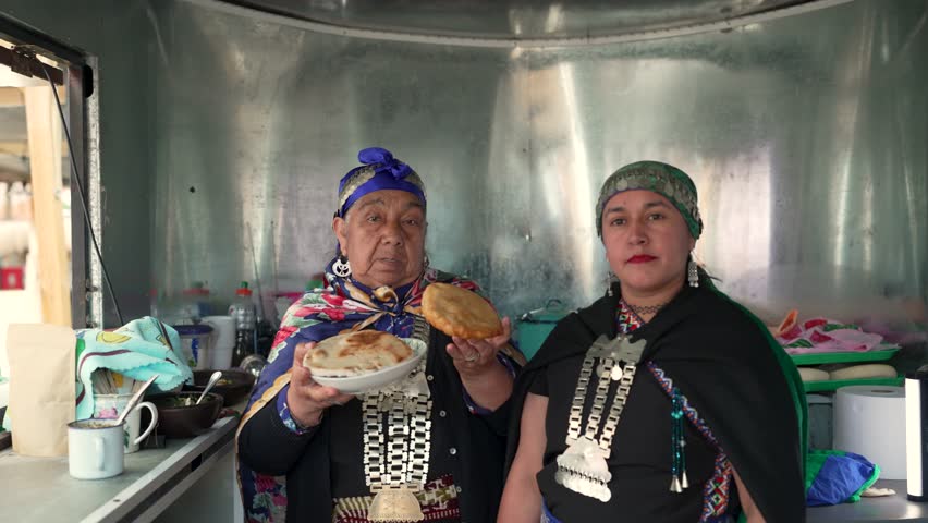 Two mapuche women in traditional clothing proudly showing and preparing typical chilean street food