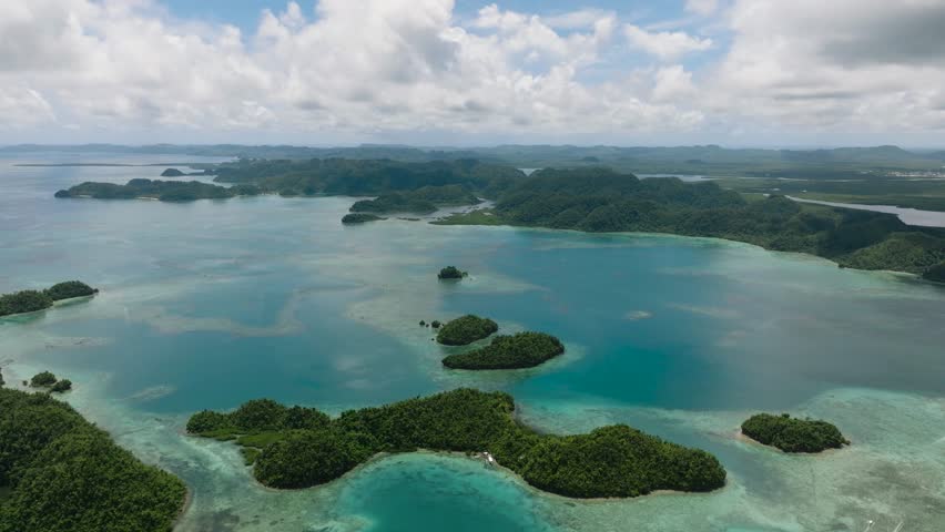 Scattered green islets lie on clear blue water with distant hills under blue sky. Sugba Blue Lagoon, Siargao, Philippines.
