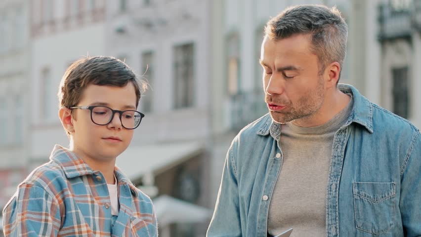Caucasian father and son using smartphone on street. Young boy with glasses learning from adult male showing mobile screen. Two generations bonding while talking outdoors in city center.