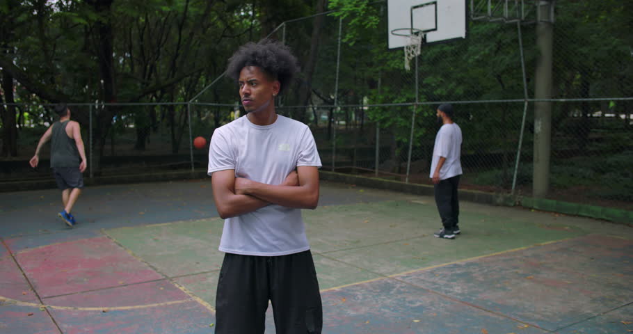 African American man standing with arms crossed on basketball court, calm focused expression while teammates play in background, outdoor urban sports setting