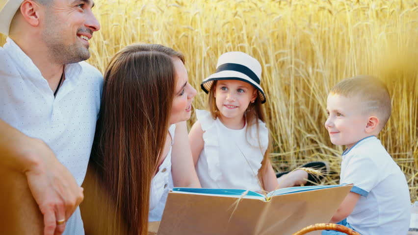 Smiling family enjoys reading book while sitting closely together in golden wheat field on sunny day, with children attentively listening