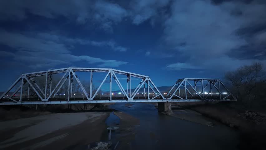 Passing train lights up this old metal bridge as it crosses over a river at night.  Night timelapse during a full moon illuminates the surrounding landscape as interstate traffic goes by in background