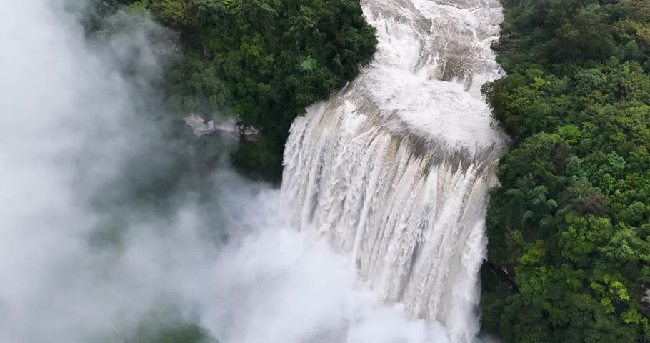 Aerial video of Huangguoshu Waterfall cascading powerfully, surrounded by lush green forest, mist rising from the base,  Guizhou Province, China.