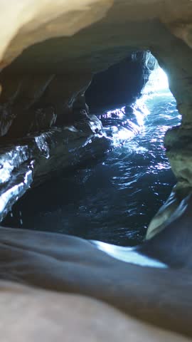 Waves flow through a narrow sea cave carved into layered rock, with sunlight illuminating the cave opening and reflecting on the moving water, creating a dramatic natural coastal scene.