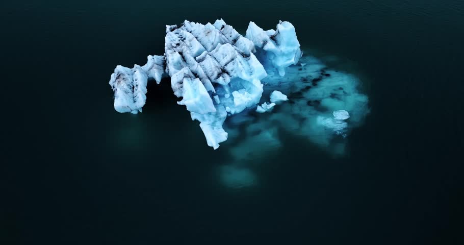 Wide overhead drone shot of a lone iceberg melting in a dark lagoon, creating a turquoise underwater plume and gentle ripples in moody overcast light.