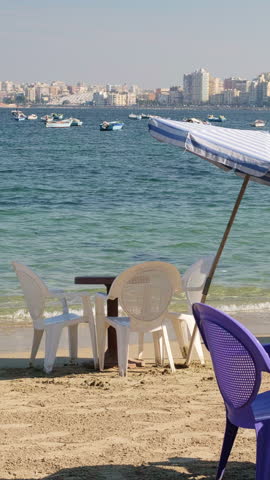 Casual plastic chairs and table arranged on sandy beach beneath striped umbrellas, overlooking calm bay with small boats and distant urban skyline on a sunny day. Harbor of Alexandria, Egypt