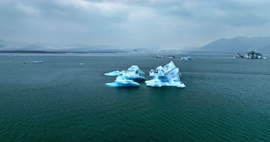 Wide drone view of two blue icebergs drifting on a calm glacial lagoon with distant mountains and an overcast sky, perfect for polar travel and climate-themed edits.