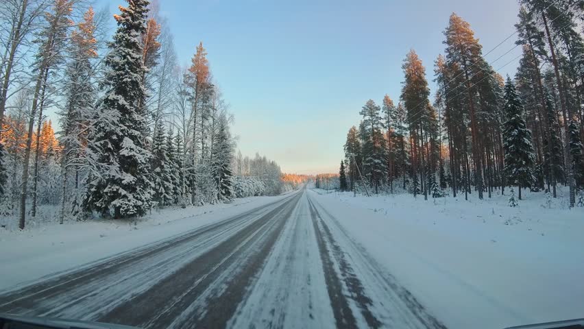Front window point of view driving on snow covered winter road in Lapland with frozen forest sunrise light icy asphalt and Nordic countryside winter travel atmosphere