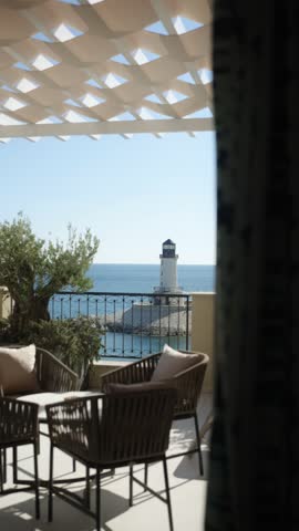 A coastal estate in Montenegro is shown from a balcony. The view includes the sea and a lighthouse under a clear sky. Seating is set up on the balcony for relaxation.