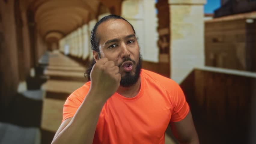 Man raising clenched fist toward camera on old town stone street with colonnade building, speaking firmly; defiance resilience.