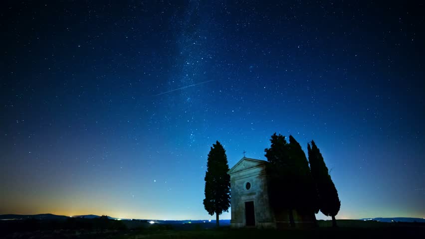 This 4K long-exposure video features star trails over a lonely chapel in Tuscany, Italy. The visual documents a stone structure flanked by silhouetted cypress trees under a night sky. The composition highlights curved light trails formed by Earth’s rotation, including a circular pattern indicating the celestial pole, capturing the intersection of historical architecture and astronomical motion in a rural landscape.
