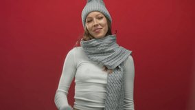 Woman points finger up and shows open palm while wearing gray knit hat scarf and glove in red studio; playful confidence. - Powered by Shutterstock - Get 15% off with code: PIKWIZARD15
