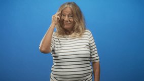 Senior woman touching temple with index finger and then pointing forward while smiling, posed in a blue studio portrait; thoughtful memory. - Powered by Shutterstock - Get 15% off with code: PIKWIZARD15