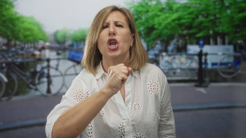 Woman with clenched fist and intense expression beckons on a canal street lined with green trees; frustration.