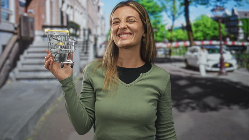 Woman holding a small shopping cart on a city street, hand raised and smiling with squinted eyes while wearing a green sweater; shopping humor joy.
