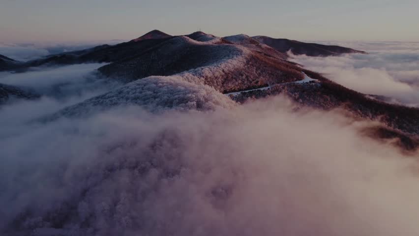 "A breathtaking aerial view of snow-dusted mountain ridges emerging from a thick sea of clouds during a serene, golden hour sunset."