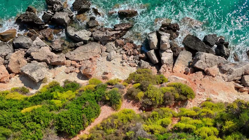 Aerial drone footage of the rugged coastline at Xigia Sulfur Beach in Zakynthos, Greece. The video shows waves crashing against large rocks and boulders along the shoreline, with clear turquoise water and greenery-covered land. Overhead flying view of the scenic landscape.