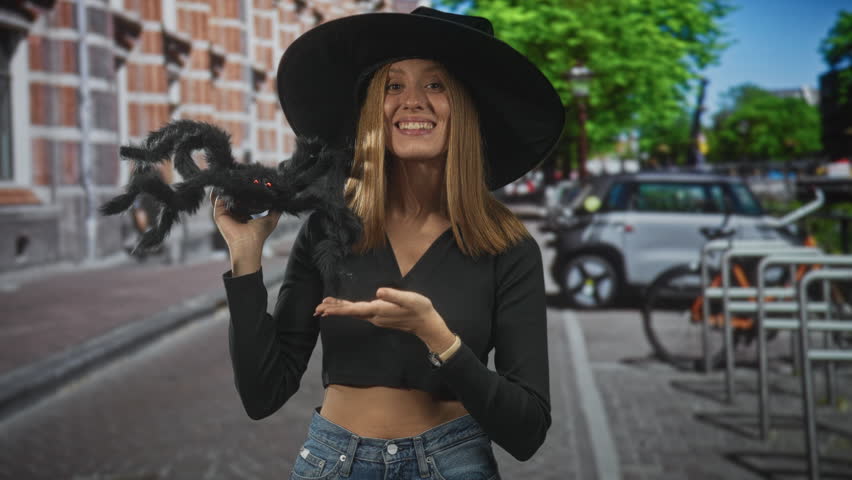Woman holding a fuzzy spider toy with open mouth, stomach exposed, on a city street; playful surprise.