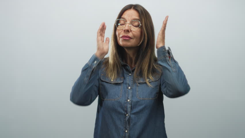 Woman covering ears with hands, pressing palms to sides of head and puckering lips in studio wearing denim shirt and glasses; refusal annoyance.