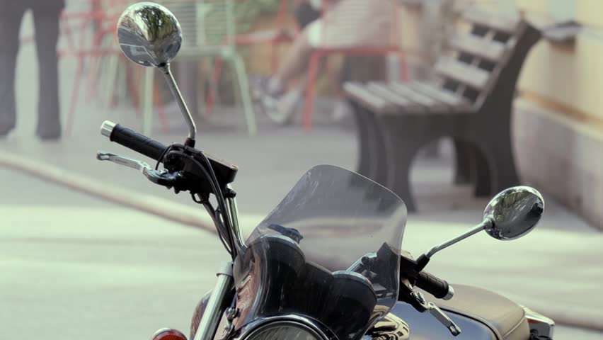 Closeup of motorcycle handlebars with mirrors and windscreen. Bench and chairs visible in blurred background