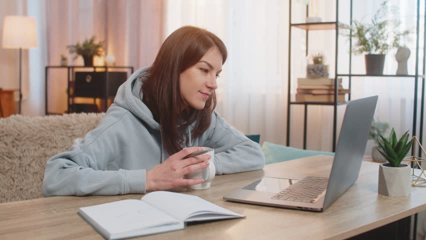 Happy young Caucasian woman freelancer drinking coffee or tea sits at table on couch looking at laptop screen working, appearing relaxed with notebook with notes lies nearby. Adult girl in home room.