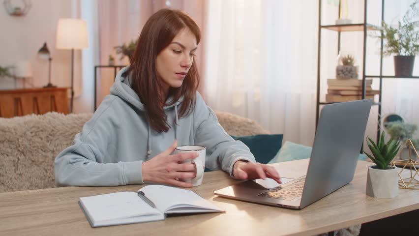 Happy young Caucasian woman freelancer drinking coffee or tea sits at table on couch looking at laptop screen working, appearing relaxed with notebook with notes lies nearby. Adult girl in home room.