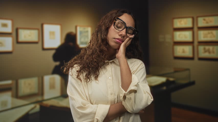 Woman with hands pressed to cheek for sleep in art gallery building, eyes closed, wearing glasses; quiet contemplation serenity.