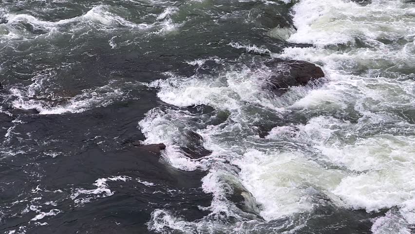 Fast moving river water rushing over large dark rocks creating strong white rapids.