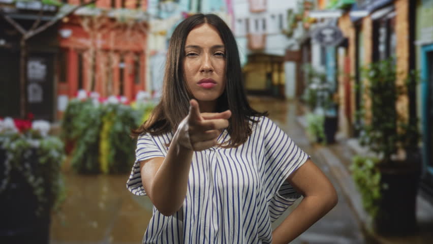Hispanic young brunette woman pointing finger directly at camera in a narrow cobbled street with potted plants and storefronts; confrontation.