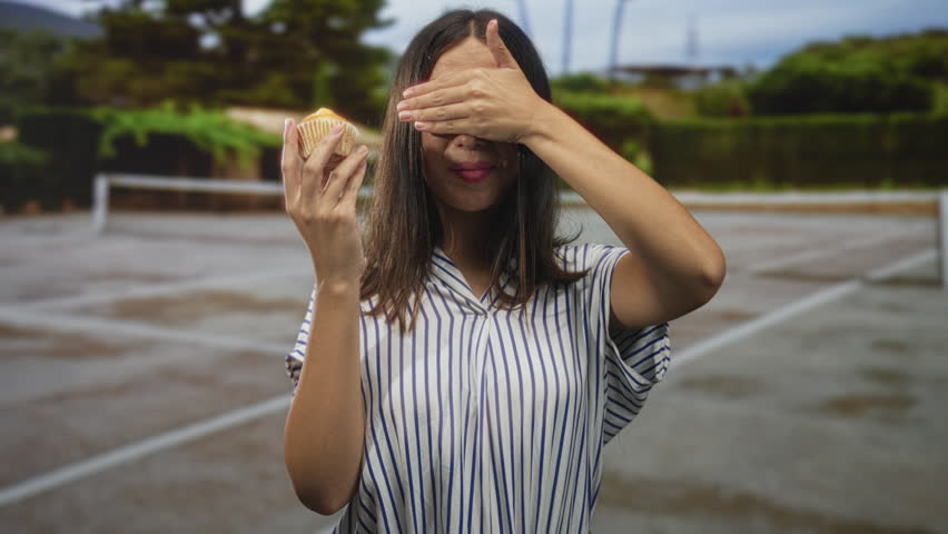 Young hispanic woman holding cupcake covering eyes with hand on tennis court beside building; playful surprise.