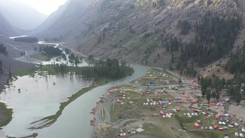 Wide aerial view of a river flowing through a mountain valley in Northern Pakistan, with small wooden cabins and tents positioned along the riverbanks, surrounded by green natural landscape