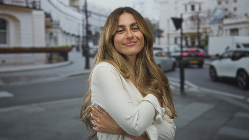 Woman with bare arms hugging herself and tousling long blonde hair while smiling softly on a busy city street; confidence comfort.