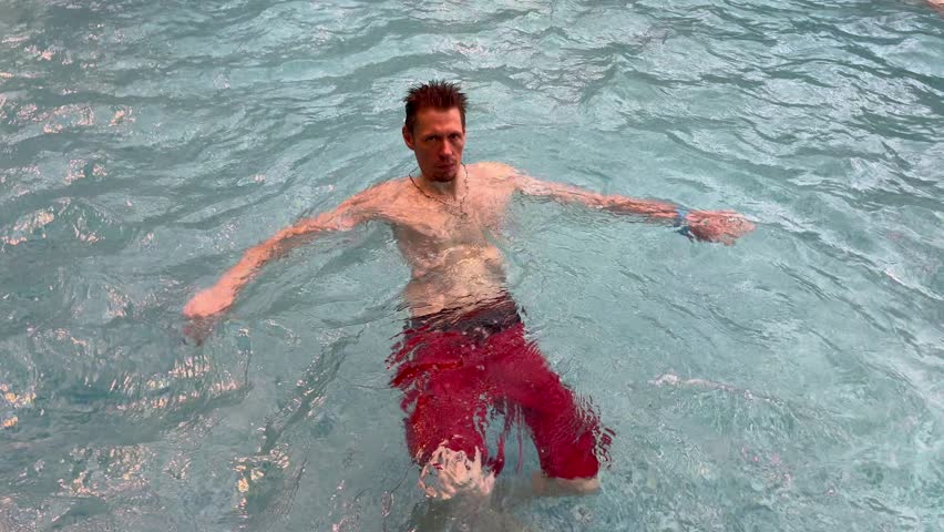 A young Caucasian man floats in a swimming pool. He has short dark hair and is wearing red swim shorts. The water is clear and blue.