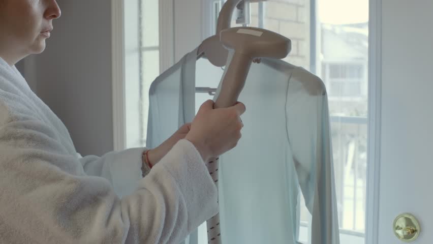 A young adult woman is seen performing household tasks by steaming a silk cardigan with a steamer in her personal room setting.