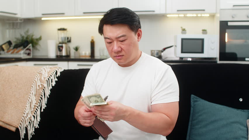 Asian man at home opens wallet sees few small dollar bills frowns sighs and sets new saving goal. Chinese guy on sofa plans budget writes note sets spend limit for week cuts costs and hopes for calm