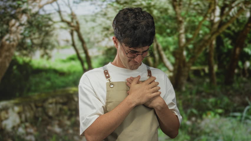 Young hispanic man wearing apron and round glasses clutching hands to chest in green park with trees and stone wall visible; vulnerability pain.