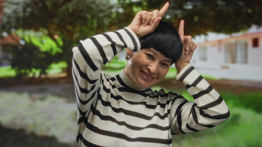 Woman smiling in an outdoor park setting with greenery, playfully posing with hands making bunny ears in vibrant daylight.
