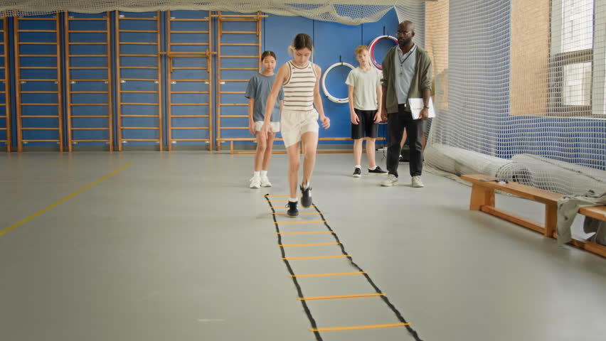 Front view shot of group of children doing agility ladder drills in indoor gym and moving towards camera one by one following each other in row, copy space