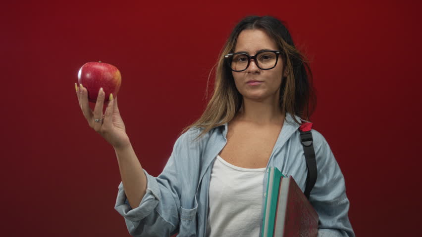 Hispanic teen woman wearing glasses and backpack holding apple and textbooks in red studio; youthful confidence.