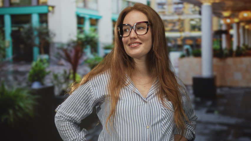 Woman wearing striped shirt and large glasses stands with hands on hips near potted plant and column by building shop in street; confidence.