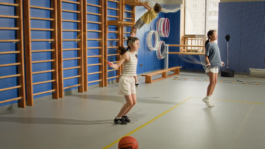 Wide angle shot of two children doing jumping rope exercises in indoor gym during PE lesson, various sports equipment in background, copy space