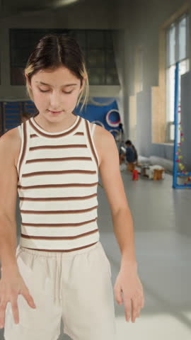 Vertical waist up shot of smiling young girl holding basketball ball and looking at camera while posing at indoor gym during sports practice or PE lesson