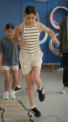 Vertical shot of children jumping on one leg towards camera one by one while doing agility exercises at indoor gym