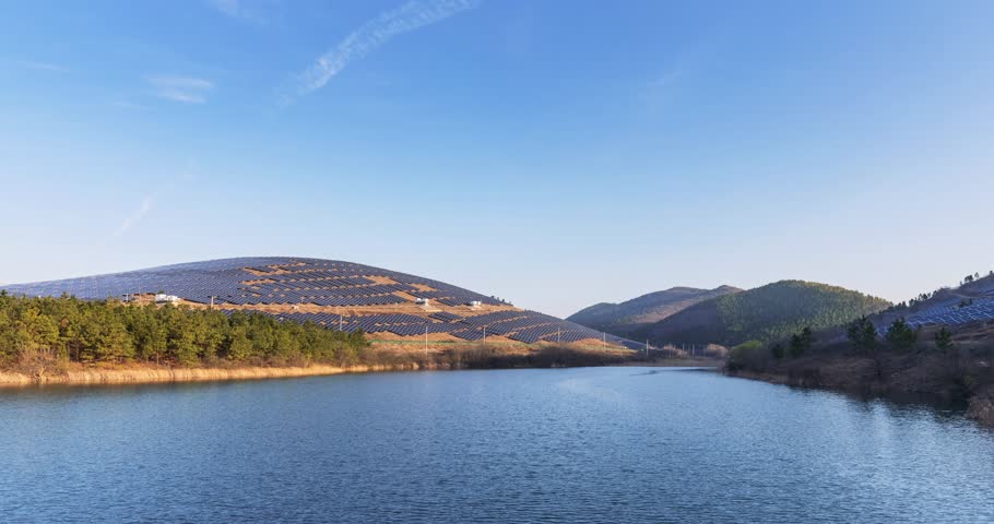 Time-lapse of a mountain solar farm at early morning, photovoltaic panel arrays on hillsides beside a calm reservoir under clear blue sky.