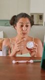 Female content creator sitting at a table filming a skincare tutorial on her smartphone, applying moisturizing cream to her face while talking to her online audience from home - Powered by Shutterstock - Get 15% off with code: PIKWIZARD15